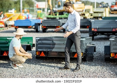 Agronomist With Salesman Choosing A New Farm Truck Trailer, Standing On The Open Ground Of The Agricultural Shop