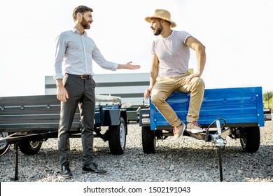 Agronomist With Salesman Choosing A New Farm Truck Trailer, Standing On The Open Ground Of The Agricultural Shop