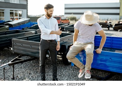 Agronomist With Salesman Choosing A New Farm Truck Trailer, Standing On The Open Ground Of The Agricultural Shop