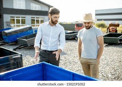 Agronomist With Salesman Choosing A New Farm Truck Trailer, Standing On The Open Ground Of The Agricultural Shop