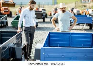 Agronomist With Salesman Choosing A New Farm Truck Trailer, Standing On The Open Ground Of The Agricultural Shop