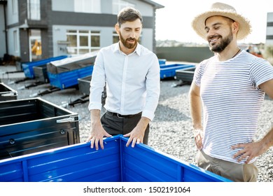 Agronomist With Salesman Choosing A New Farm Truck Trailer, Standing On The Open Ground Of The Agricultural Shop