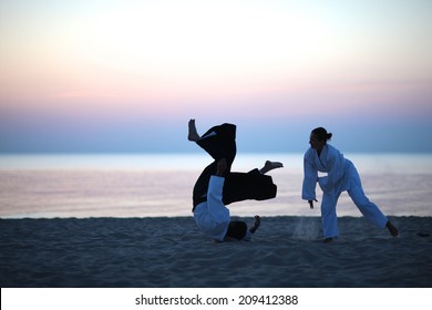 Aikido practice on the beach at sunset; student in white kimono throwing aikido master 