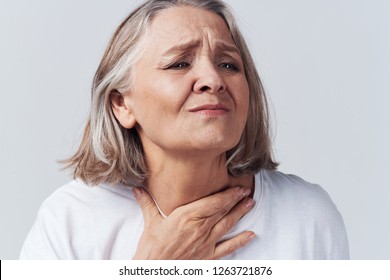 Ailing elderly woman with a sore throat holding on to her neck against a gray background                           