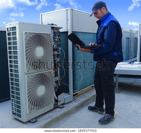 air conditioning technician\
making a diagnosis of an industrial air conditioning unit with a\
laptop next to other VRV condenser units on a rooftop in a sunny\
day