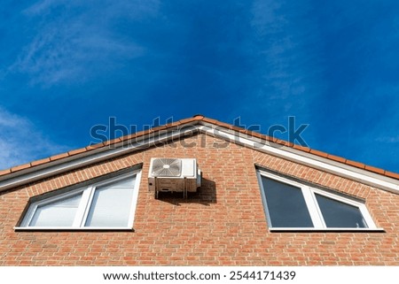 An air conditioning unit is fixed to the side of a modern brick house, with bright blue skies and wispy clouds above, highlighting the architectural features.