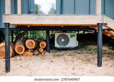 An air conditioning unit is installed under a wooden platform, positioned next to cut logs on sandy terrain. The environment suggests a rural setting where the structure blends with natural elements.