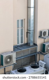 Air conditioning units and a vertical ventilation duct on a pale beige building exterior, showing metal condensers, fans, pipes and louvers urban HVAC infrastructure with an industrial utilitarian aesthetic.