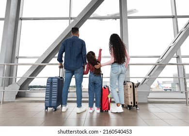 Air Journey. Family With Daughter Looking At Plane Departure Out Of Window In Airport, Little Girl Pointing Away, Rear View Shot Of Young Parents And Female Child Standing With Suitcases At Terminal