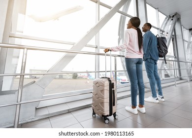 Air Travel. Black Millennial Couple Standing Together Near Window In Airport, Looking At Flying Plane, Happy African American Man And Woman With Luggage Waiting For Flight Boarding, Low Angle View