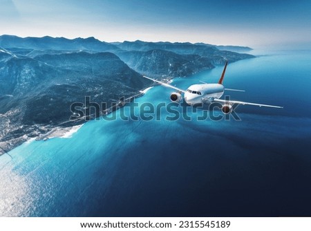 Aircraft is flying over islands and sea at sunrise in summer. Landscape with white passenger airplane, seashore, mountains, forest, clear sky, and blue water in bright day. Travel and resort. Tourism