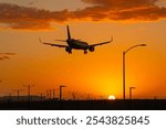 An aircraft landing at Los Angeles Airport at sunset, orange hazy sky with orange colored clouds. Airport infrastructure only visible as dark silhouettes.