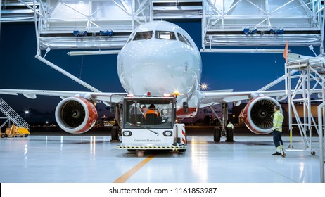 Aircraft Maintenance Hangar Where New Airplane Is Toed By A Pushback Tractor/ Tug Onto Landing Strip. Crew Of Mechanics, Engineers And Drivers Works Busily.