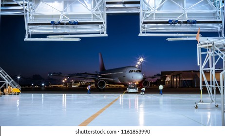 Aircraft Maintenance Hangar Where New Airplane Is Toed By A Pushback Tractor/ Tug Onto Landing Strip. Crew Of Mechanics, Engineers And Drivers Works Busily.