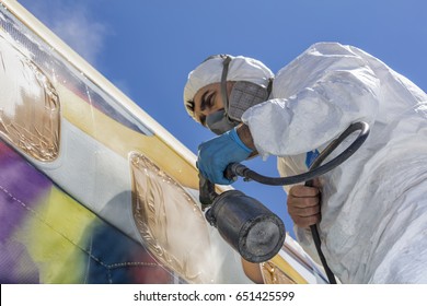 Aircraft Painting And Sandblasting.  Spray Painting, Airplane Wing. Worker In Coverall Using Airbrush. Passenger Aircraft During Maintenance And Painting.