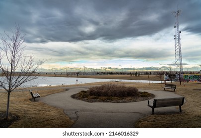 Airdrie Alberta Canada, April 04 2022: A Frozen Storm Water Retention Pond And Park Benches At An Off Leash Dog Park With A Distant Communications Tower.