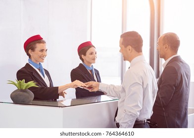 Airline Passengers Checking In At Airline Counter. Young Woman Giving Passport And Ticket Back To Businessman At Airport Check In. Two Businessmen At Airport Checkin Desk Leaving On Business Trip.