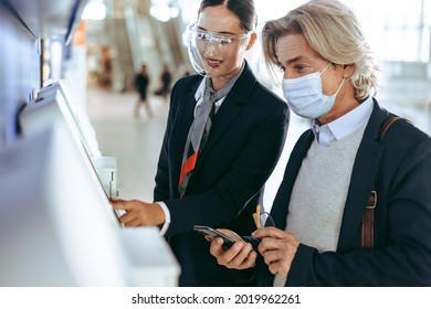 Airline Staff Helping A Business Traveler In Doing The Self Check-in At The Airport Terminal During Covid-19 Pandemic. Masked Businessman At Self Service Check In Machine With Flight Attendant.