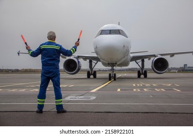 An airliner is taxiing into a parking lot at an airport, an air marshall with batons indicates a parking spot, an aeronautical engineer helps pilots park the plane