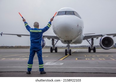 An airliner is taxiing into a parking lot at an airport, an air marshall with batons indicates a parking spot, an aeronautical engineer helps pilots park the plane
