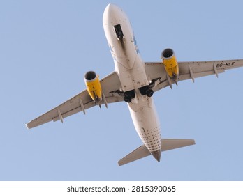 Airplane in flight viewed from below, featuring yellow engines and visible landing gear. the aircraft is seen against a blue sky