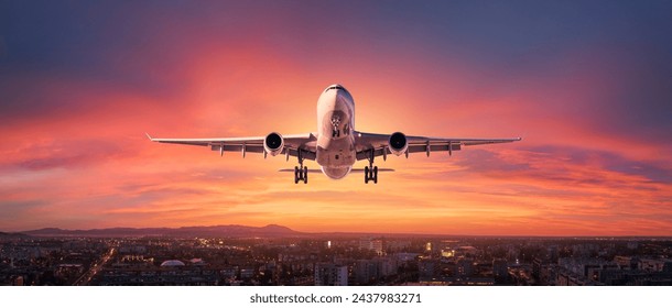 Airplane is flying in colorful sky over city at sunset. Landscape with passenger airplane, skyline, purple sky with red and pink clouds at dusk. Aircraft is landing at twilight. Aerial view of plane - Powered by Shutterstock