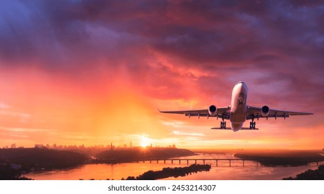 Airplane is flying in colorful sky over city at sunset. Landscape with passenger airplane, skyline, purple sky with red and pink clouds at dusk. Aircraft is landing at twilight. Aerial view of plane - Powered by Shutterstock