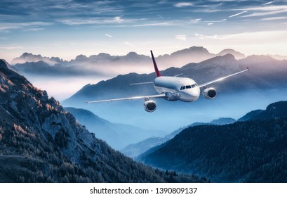 Airplane is flying over mountains in fog at sunset in summer. Landscape with passenger airplane, hills in low clouds, blue sky. White aircraft. Business travel. Commercial plane. Aerial view - Powered by Shutterstock