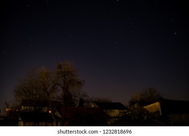 Airplane Leaving Light Trail While Flying Over Town Late At Night. Stars Shining Bright Over Town In Autumn Night