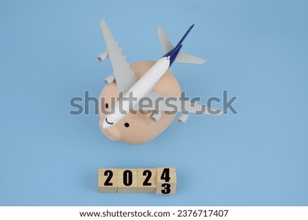 Airplane on piggy bank and wooden cubes with 2023 and 2024 on blue background. Save money and airline tickets prices in year 2024.