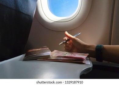 Airplane Tray Table On Seat Back Inside Cabin; One Hundred Thousand Rupiahs, Indonesian Currency And Customs Declaration Form With Sun Shining Through The Window. Travel Lifestyle Vacations Concept.