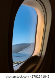 Airplane window frames clouds and wing during a calm evening flight. Soft sunset colors spread across the sky above the horizon.