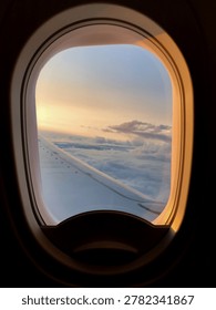 Airplane window frames clouds and wing during a calm evening flight. Soft sunset colors spread across the sky above the horizon.