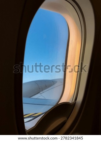 Airplane window frames clouds and wing during a calm evening flight. Soft sunset colors spread across the sky above the horizon.