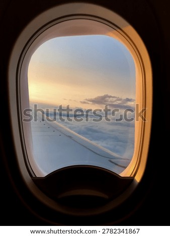 Airplane window frames clouds and wing during a calm evening flight. Soft sunset colors spread across the sky above the horizon.