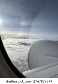 Airplane wing and engine viewed through window above fluffy clouds, with bright sunlight and a faint halo shining in the sky.