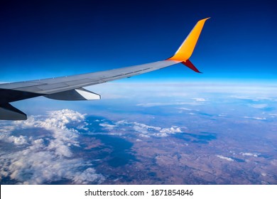 Airplane Wing Flying Over White Clouds And Brown Earth In A Blue Atmosphere. Aerial View Of The Planet From A Plane Window