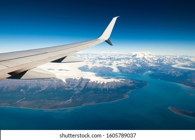 Airplane wing view with snow capped mountains in a cold environment. On board a Milk Run flight, between two small cities in the state of Alaska with a beautiful natural landscape in sight. - Powered by Shutterstock