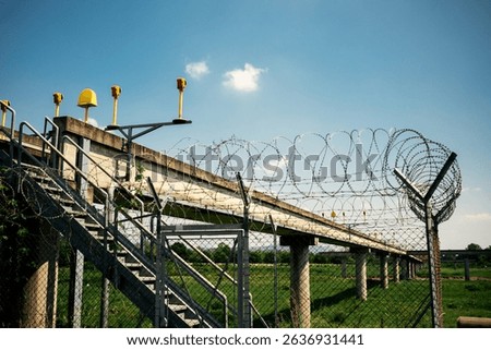 Airport approach lighting structure with barbed wire fence and access stairs under clear blue sky, perimeter security installation.