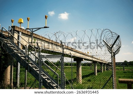 Airport approach lighting structure with barbed wire fence and access stairs under clear blue sky, perimeter security installation.