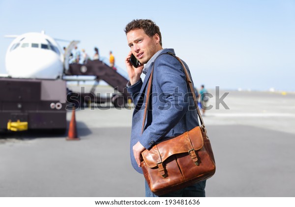 Airport business man on smartphone by plane. Young\
male professional hip businessman talking on smartphone boarding\
airplane going flying on business trip. Casual male wearing suit\
and laptop bag.