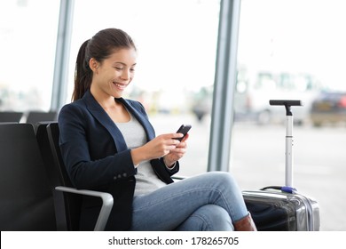 Airport Business Woman On Smart Phone At Gate Waiting In Terminal. Air Travel Concept With Young Casual Businesswoman Sitting With Hand Luggage Suitcase. Beautiful Young Mixed Race Female Professional