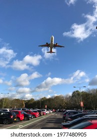 Airport Car Park With Jet Plane Overhead Taking Off. Vehicles In Parking Lot With Aeroplane Flying In Blue Sunny Sky With White Fluffy Clouds. Summer Holiday Departures, Or Business Travel