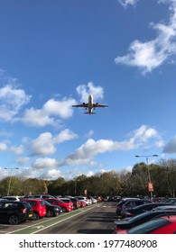 Airport Car Park With Jet Plane Overhead Taking Off. Vehicles In Parking Lot With Aeroplane Flying In Blue Sunny Sky With White Fluffy Clouds. Summer Holiday Departures, Or Business Travel