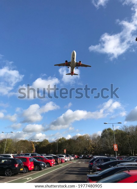Airport car park with jet plane overhead taking\
off. Vehicles in parking lot with aeroplane flying in blue sunny\
sky with white fluffy clouds. Summer holiday departures, or\
business travel