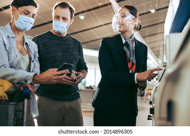 Airport Staff With Couple Helping In Filling Details In Self Service Machine During Pandemic. Tourist Couple In Face Masks At Self Service Helped By Airlines Attendant At Airport.