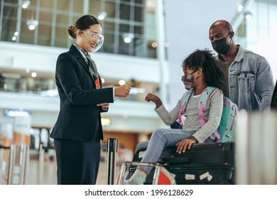 Airport Staff During Pandemic Giving Fist Bump To Young Girl Of Traveler Family At Boarding Gate. Ground Staff At Airport Greeting Girl Kid In Face Mask Sitting On Luggage.