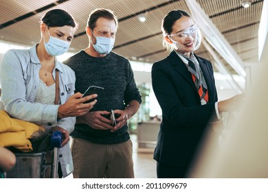 Airport Staff In Face Shield Helping Couple In Face Masks During Pandemic. Male And Female Tourists In Face Masks Helped By Ground Attendant At Airport.