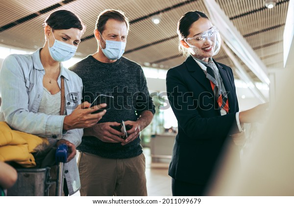 Airport staff in face shield helping couple\
in face masks during pandemic. Male and female tourists in face\
masks helped by ground attendant at\
airport.
