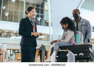 Airport Staff Standing At Boarding Gate With Girl Sitting On Luggage And Father Pushing The Trolley. Ground Attendant At Airport Giving Help To Tourist Family In Face Masks During Pandemic.